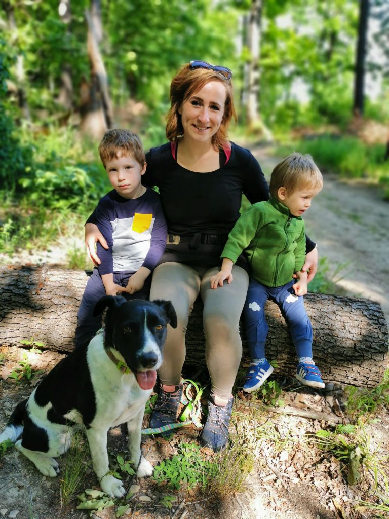 LucaKutya túrázik; anya a két fiával ül egy farönkön, előttük Luca